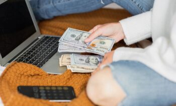 A woman sitting on her living room floor, using a laptop, and counting cash.