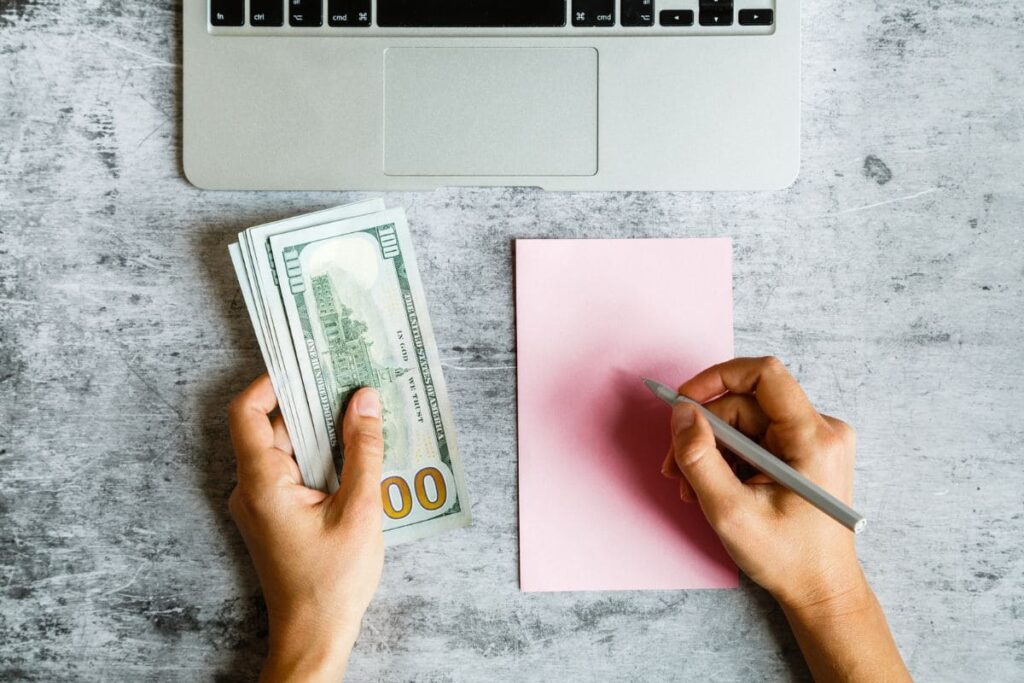 A woman sitting at a desk, using a laptop, counting cash, and writing on a notepad.