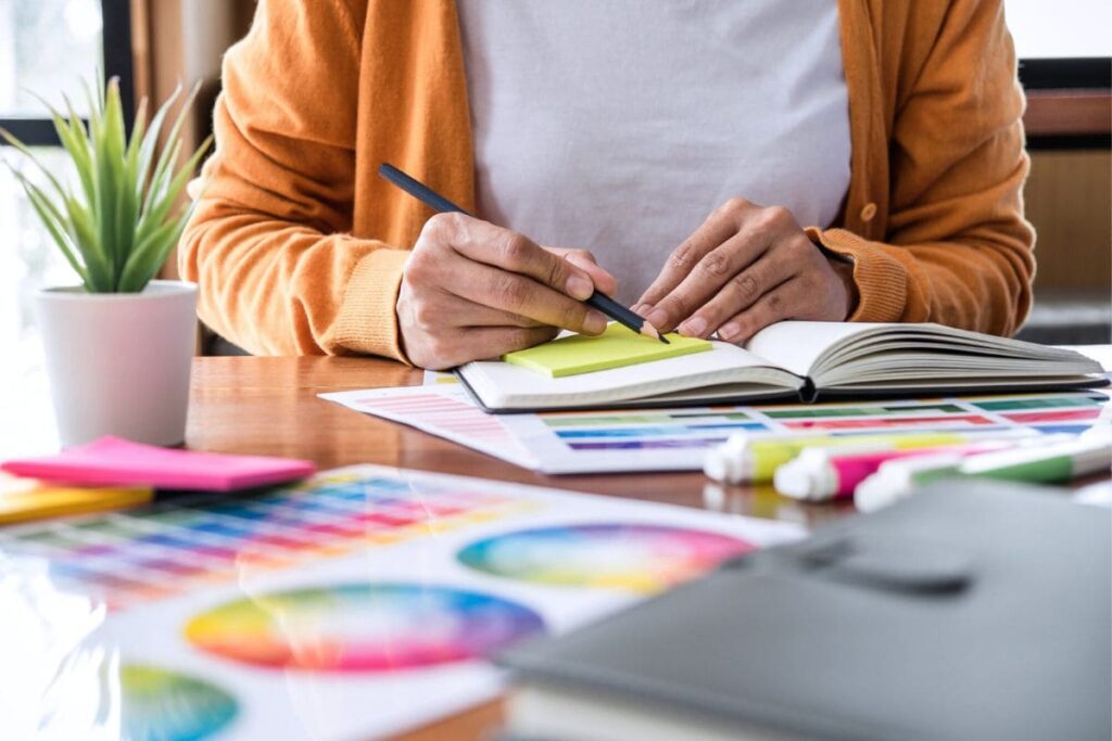 A freelance graphic designer working at her home office desk and writing in a notebook.