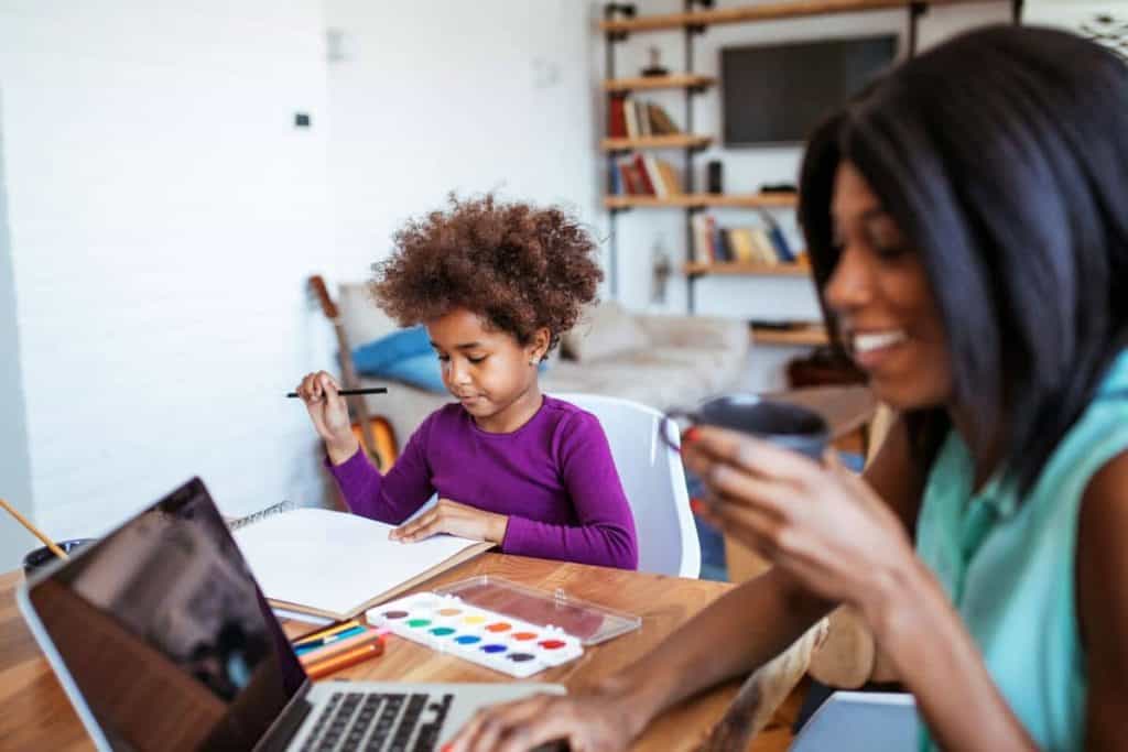 A stay-at-home mom working at the kitchen table on her laptop while her daughter draws.