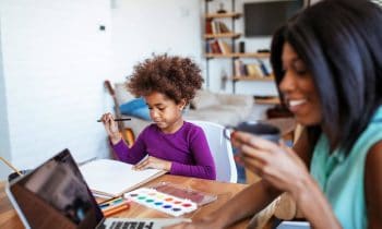 A stay-at-home mom working at the kitchen table on her laptop while her daughter draws.