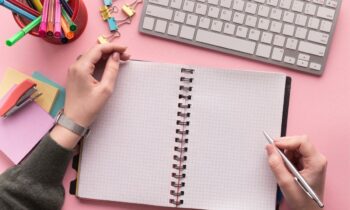 Overhead view of a woman's hands writing in a notebook while sitting at a desk.