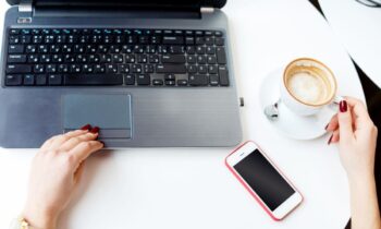 A woman working at a home office desk, with a laptop, cell phone, and coffee.