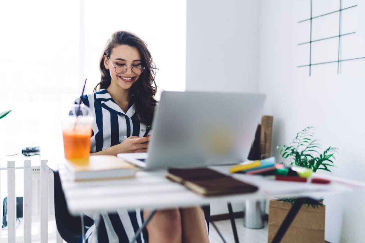 A remote freelancer is working from her home office at a desk, using a laptop and a smart phone.