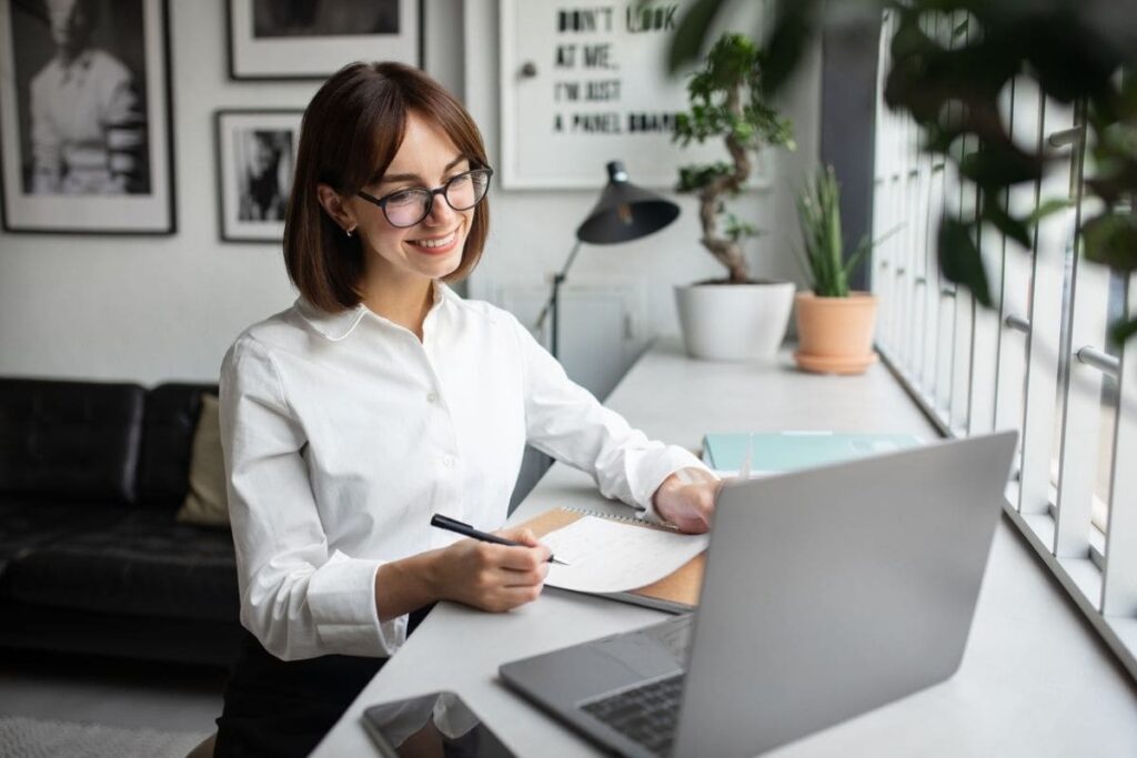 A remote freelancer is working from her home office at a desk, using a laptop and writing on a notepad.