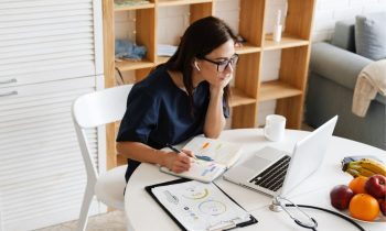 A virtual medical scribe working from home at a table, using a laptop and earphones to work.