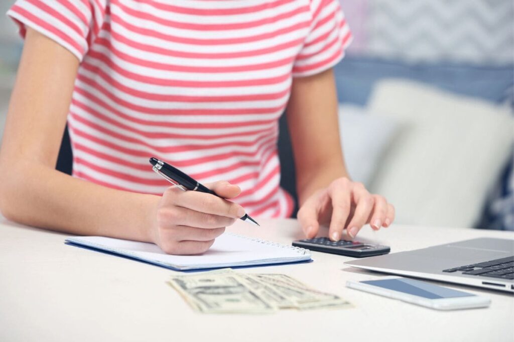 A woman sitting at her home office desk, using a calculator and counting cash to create a budget.