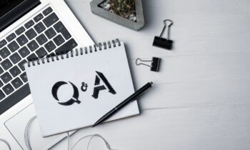 A desk with a laptop, ear phones, a cactus, and a notebook with "Q&A" written in it.