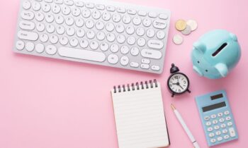 A work from home office desk with a keyboard, notebook, coins, a clock, a calculator, and a piggy bank.