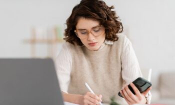 A woman at home sitting at her home office desk, using a laptop and cell phone.