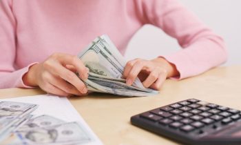 A woman sitting at a desk in her home office, counting cash for her bills and using a calculator.