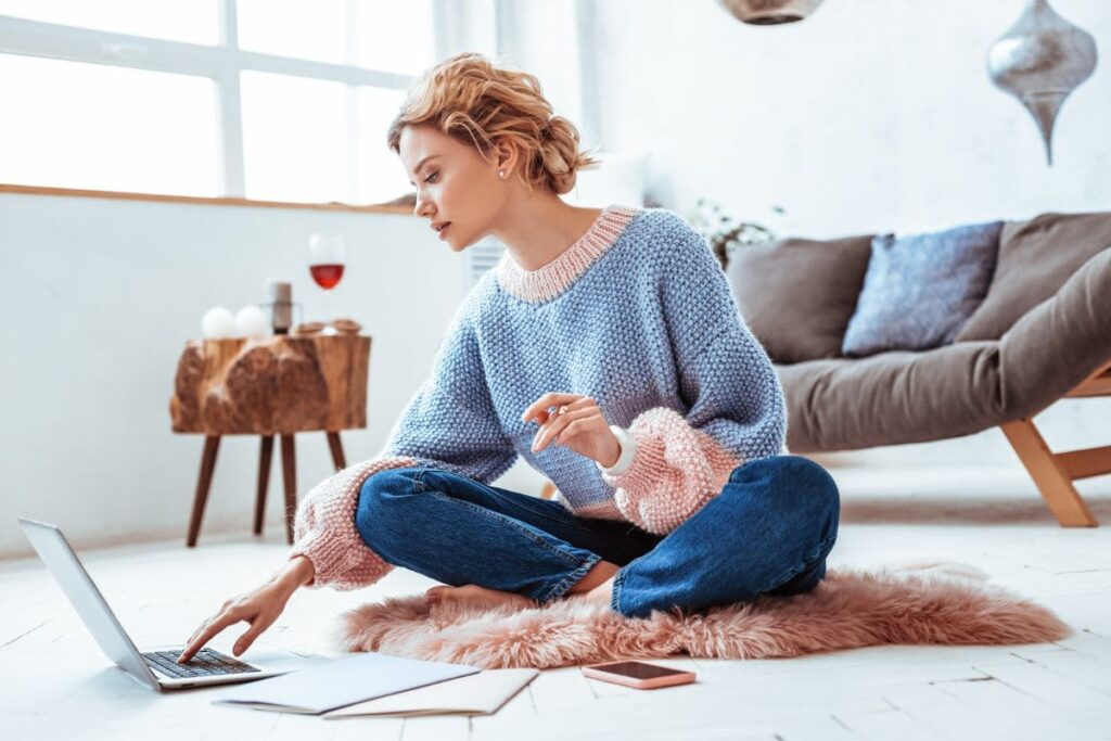 A woman sitting cross-legged on her living room floor, working from home on a laptop.