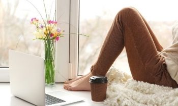 Woman sitting at home on window sill, working on laptop.