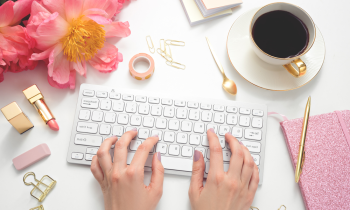 woman sitting at desk typing on keyboard, sitting on her desk is coffee, flowers, lipstick, a pen and notebook