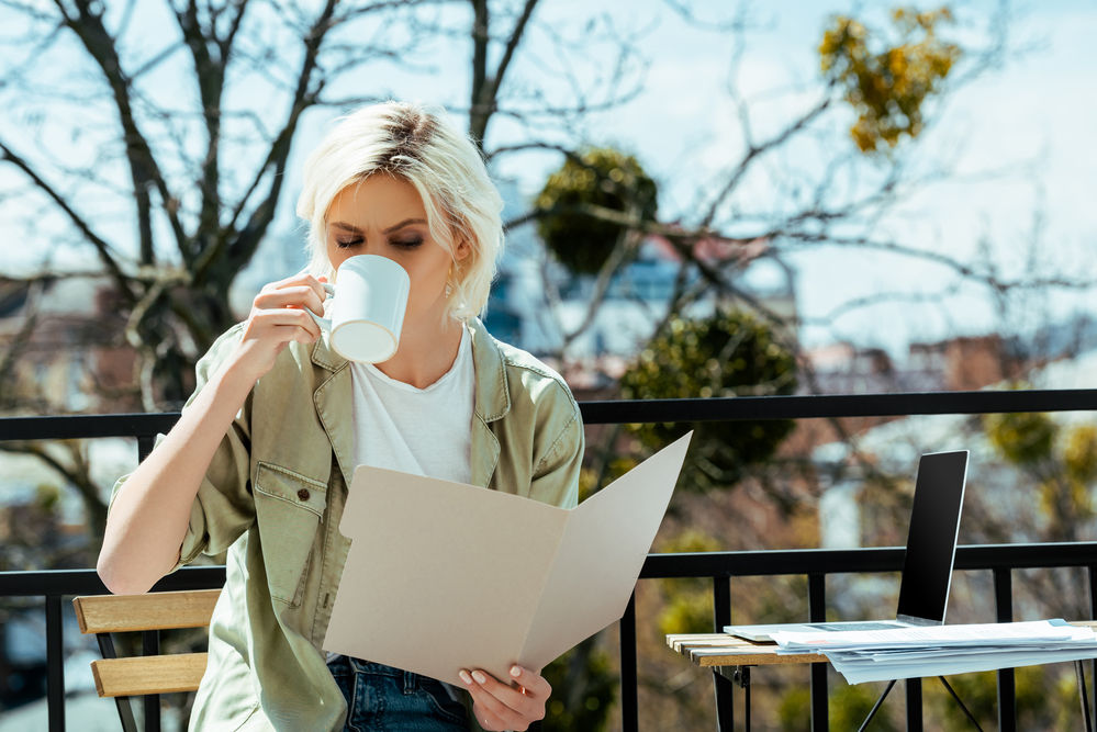 woman with blonde hair outdoors drinking coffee and reading a file folder with documents for her online side job